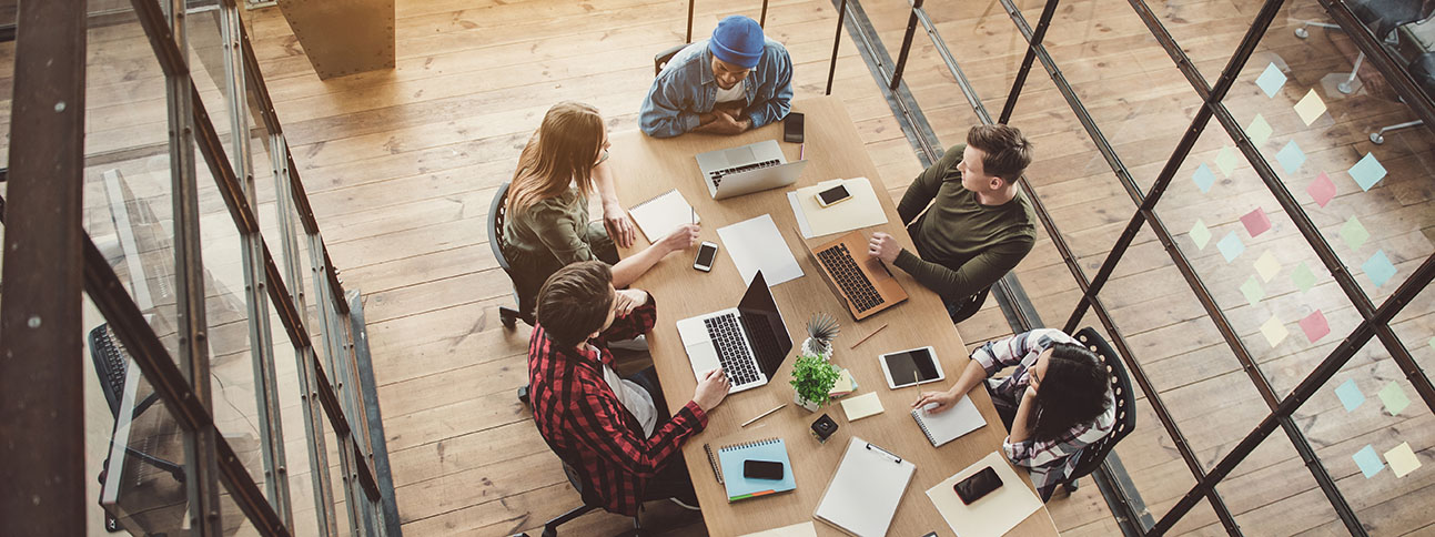 a group of five people are gathered around a table with laptops, mobile devices, and pencils engaged in a lively discussion probably about software development