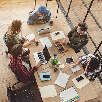 a group of five people are gathered around a table with laptops, mobile devices, and pencils engaged in a lively discussion probably about software development