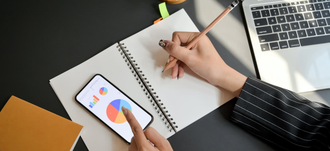 Woman writing on notebook and looking at charts on cell phone