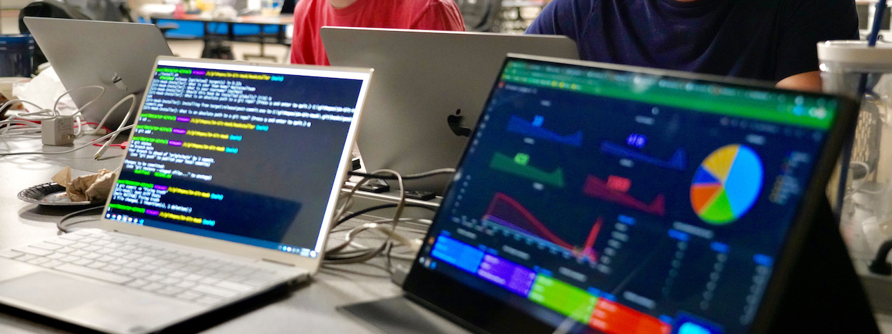 A chaotic workspace featuring four computers, one showing a terminal window and another showing a visualization dashboard.