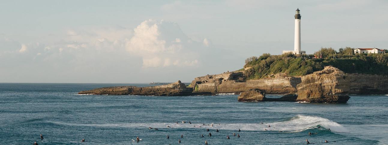 A lighthouse on a cliff off in the distance, surrounded by waves and birds.