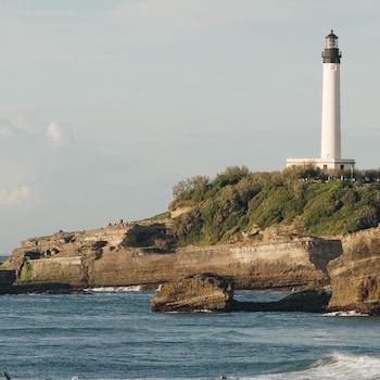 A lighthouse on a cliff off in the distance, surrounded by waves and birds.