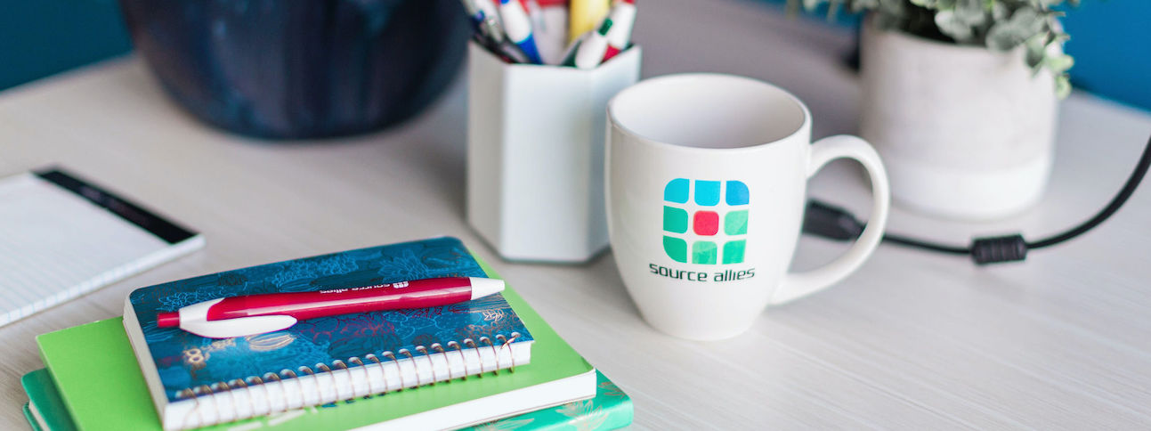 An inviting desk space complete with a white coffee mug with the Source Allies logo on it, a plant, a stack of notebooks, and a pen.