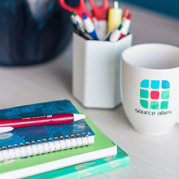 An inviting desk space complete with a white coffee mug with the Source Allies logo on it, a plant, a stack of notebooks, and a pen.